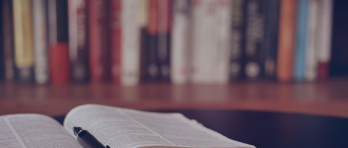 closeup of an open book and pen with several books on a shelf behind